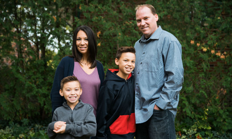 Family of a dark-haired mom, tall dad, and two dark-haired young boys smiling in a green forest wearing longs sleeves
