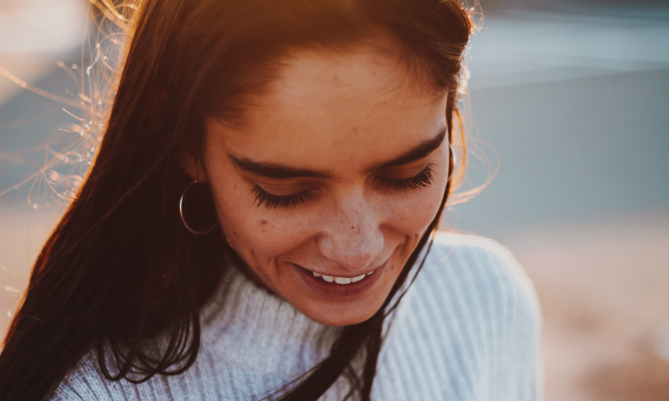 Brunette woman with white sweater and silver hoop earrings looks down and smiles with her restored smile as the sun sets