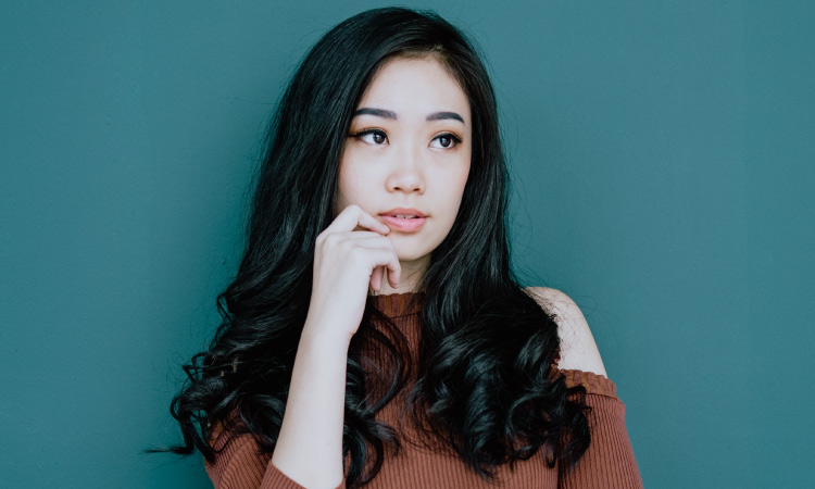 Brunette woman wearing a rust-colored sweater puts her hand ponderously to her chin and looks up diagonally