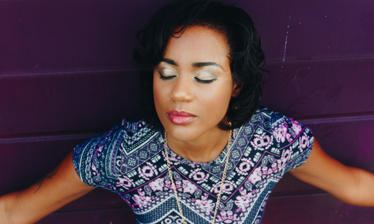 Dark-haired woman wearing a purple patterned blouse leans against a purple wall with her eyes closed, trying to relax