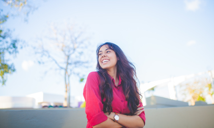Brunette young woman wearing a red blouse and a watch smiles and looks to the side in a parking lot on a cloudless day