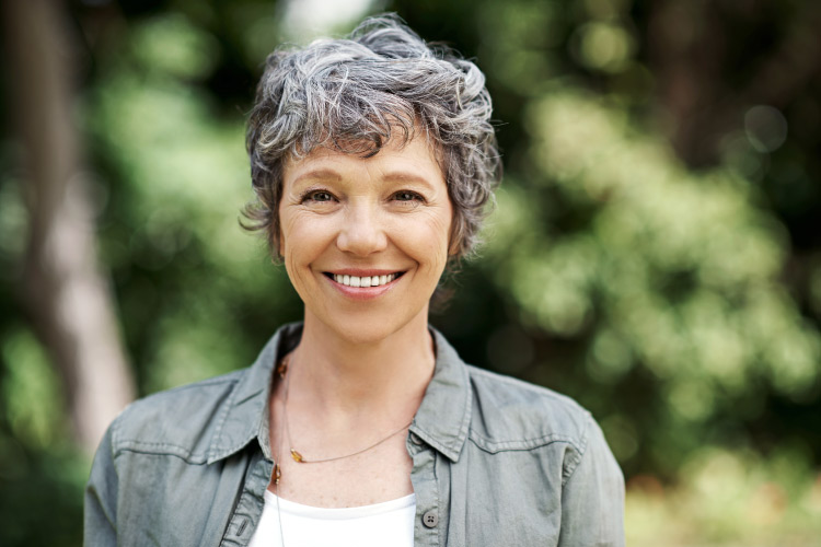 Gray-haired woman outside wearing a gray button-up over a white tanktop smiles with a natural-looking dental crown