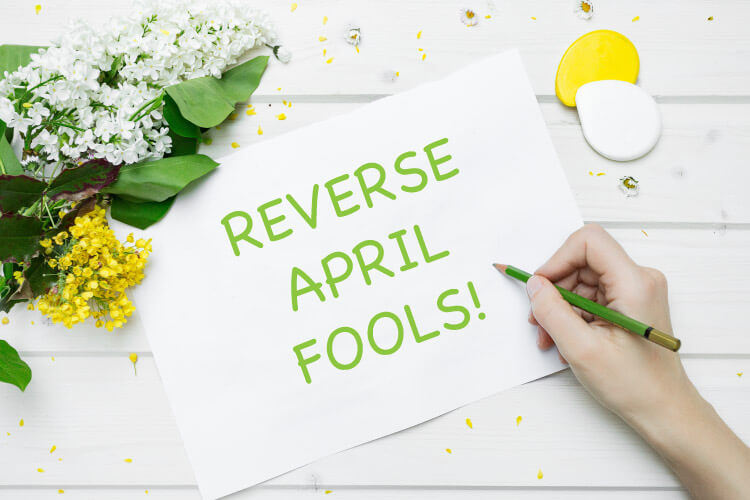 Tabletop view of a white paper with green REVERSE APRIL FOOLS next to a bunch of spring flowers and a green pencil