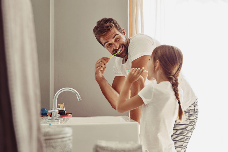 Brunette dad wearing his pajamas brushes his teeth next to his young daughter at the bathroom sink