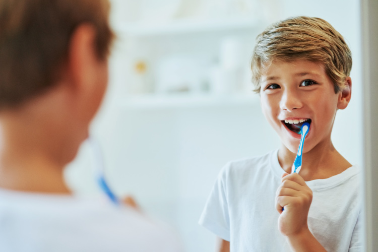 Young boy brushes his teeth at home with a blue toothbrush while smiling at his reflection