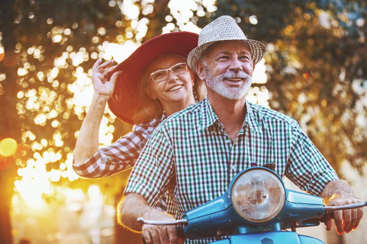 A smiling woman with a big sun hat behind a smiling man in a plaid shirt on a turquoise scooter 