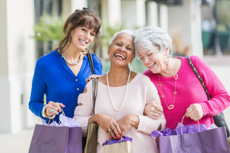 Three smiling & laughing mature ladies with arms linked walking down the street with shopping bags