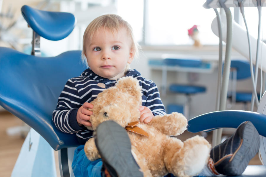 Blonde toddler boy sitting in the dental chair with his teddy bear for is first dental visit.